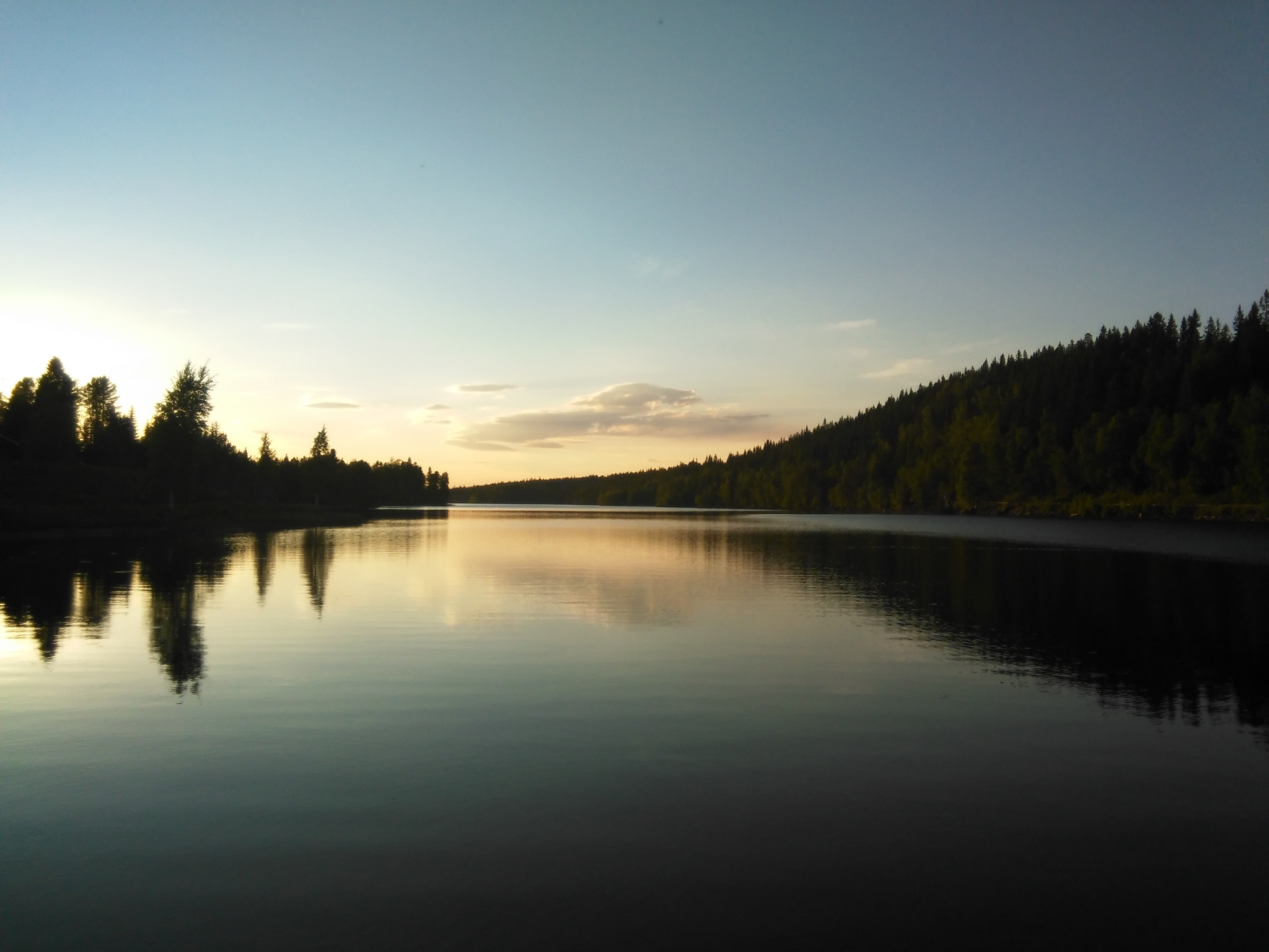 Ein Sonnenuntergang über einem See in Schweden taucht den See in buntes Licht, im See spiegeln sich die Bäume am Ufer, im Hintergrund sind einzelne Wolken